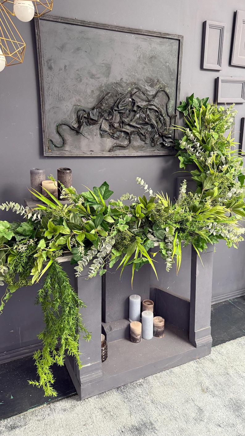 May include: A gray fireplace mantel decorated with a green garland of eucalyptus, ivy, and other greenery. The mantel has a gray background and is decorated with candles.