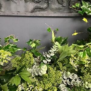 Pode incluir: Um close-up de um arranjo floral verde com folhas de eucalipto, hera e pequenas flores brancas. O arranjo est&aacute; sobre uma superf&iacute;cie de madeira branca.