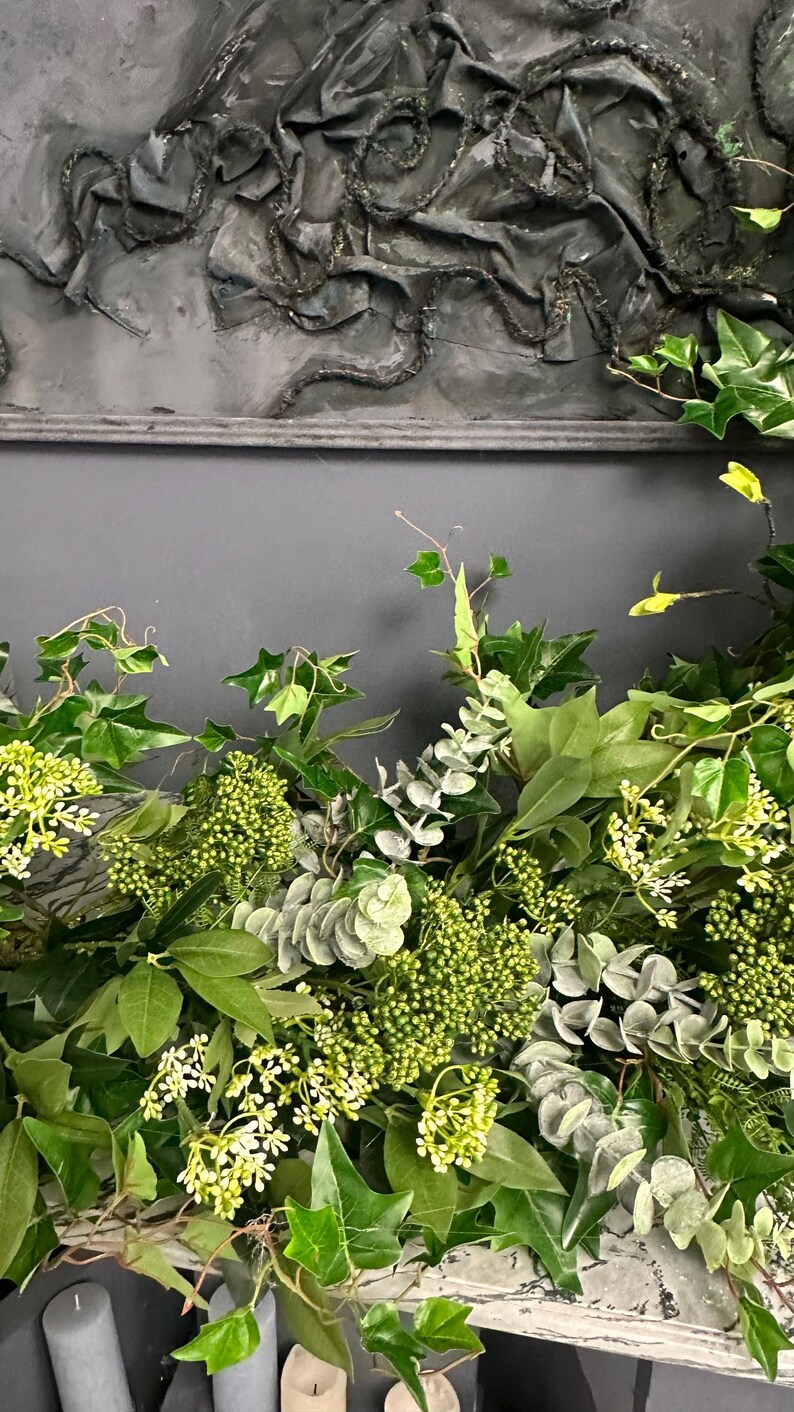 Pode incluir: Um close-up de um arranjo floral verde com folhas de eucalipto, hera e pequenas flores brancas. O arranjo est&aacute; sobre uma superf&iacute;cie de madeira branca.
