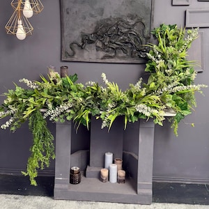 May include: A gray fireplace mantel with a green garland of leaves and flowers. There are five candles on the mantel, three are white and two are brown.