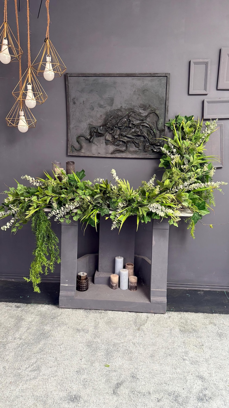 May include: A gray fireplace mantel with a green garland of leaves and flowers. There are five candles on the mantel, three are white and two are brown.