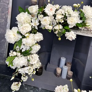 以下が含まれることがあります： 暗い灰色の表面に、白いシャクヤクやその他の白い花が滝のように配置されています。このアレンジメントには緑の葉が含まれており、さまざまなグレーとブラウンの色合いのキャンドルがアクセントになっています。装飾的なフラワーアレンジメント。