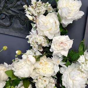 以下が含まれることがあります： 白いシャクヤクやその他の白い花と緑の葉の豪華なアレンジメント。 花は密集して配置され、完全で質感のあるディスプレイを作成します。 背景は濃い灰色です。
