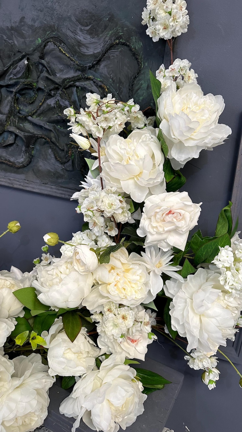 以下が含まれることがあります： 白いシャクヤクやその他の白い花と緑の葉の豪華なアレンジメント。 花は密集して配置され、完全で質感のあるディスプレイを作成します。 背景は濃い灰色です。