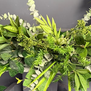 May include: A lush green garland made of various types of foliage, including eucalyptus, ferns, and other leafy plants. The garland is draped over a surface and has a natural, rustic look.