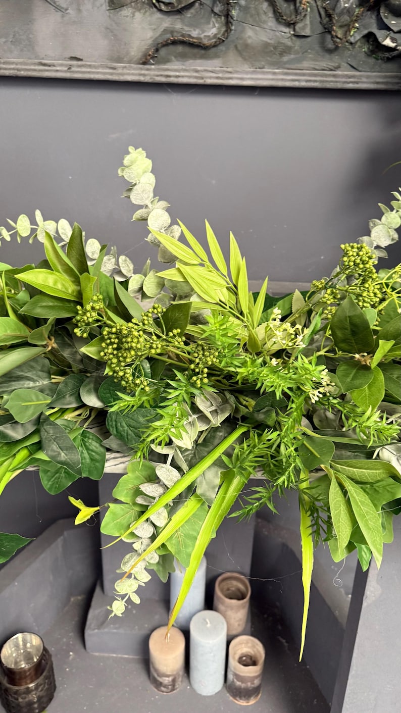 May include: A lush green garland made of various types of foliage, including eucalyptus, ferns, and other leafy plants. The garland is draped over a surface and has a natural, rustic look.