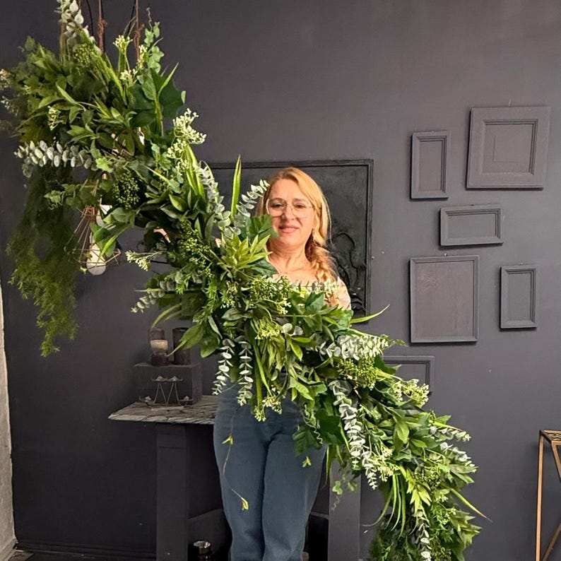 May include: A woman holds a large, lush green garland made of various types of greenery, including eucalyptus, ferns, and other leafy plants. The garland is long and flowing, and the woman is smiling as she holds it up.