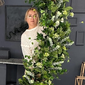 Pode incluir: Uma mulher est&aacute; em frente a um grande arranjo floral verde e exuberante. O arranjo &eacute; composto por v&aacute;rios tipos de vegeta&ccedil;&atilde;o, incluindo eucalipto, hera e samambaias. A mulher est&aacute; vestindo um su&eacute;ter branco e botas brancas.
