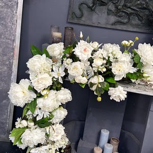 以下が含まれることがあります： 白いシャクヤクやその他の白い花と緑の葉が、濃い灰色の表面に配置されています。アレンジメントは表面の側面を滝のように流れ落ちています。背景にはいくつかのキャンドルが見えます。