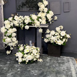 以下が含まれることがあります： 白い花、シャクヤクやバラなどを使ったアレンジメントが、濃い灰色の暖炉と周囲を飾っています。花のディスプレイには、床に大きなアレンジメントと、黒い立方体の上に小さなアレンジメントが含まれています。
