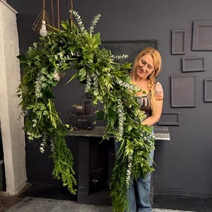May include: A woman holds a large, lush green garland made of various types of greenery, including eucalyptus and ferns. The garland is shaped like a circle and is hanging from the ceiling.
