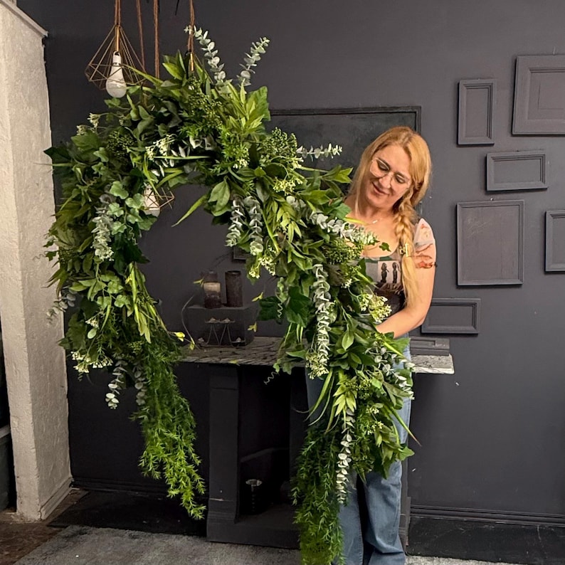 May include: A woman holds a large, lush green garland made of various types of greenery, including eucalyptus and ferns. The garland is shaped like a circle and is hanging from the ceiling.