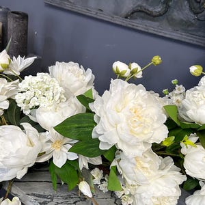 以下が含まれることがあります： 白いシャクヤクやその他の白い花と緑の葉のディスプレイ。 花は、グレーと白の大理石模様の表面に配置されています。 背景には、濃い灰色の壁と装飾品が含まれています。