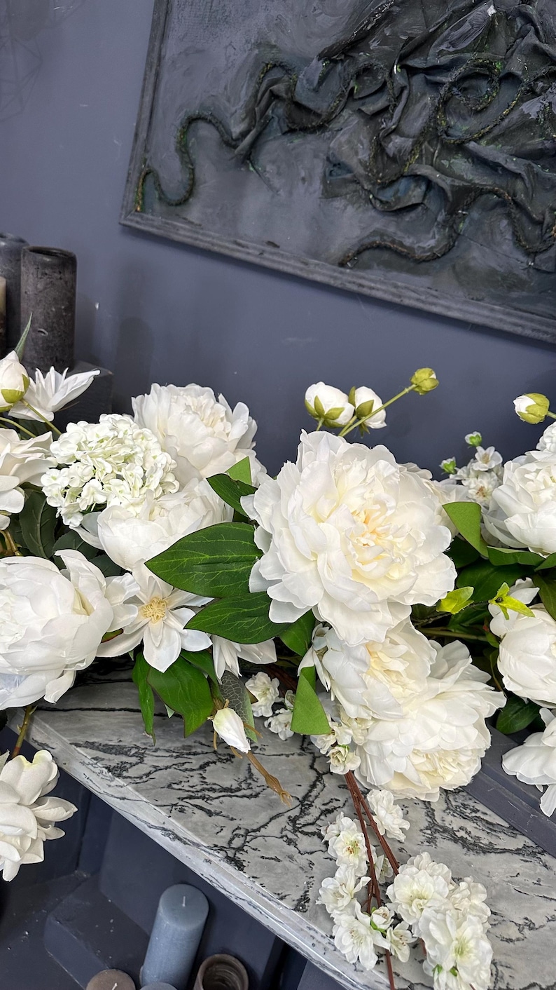 以下が含まれることがあります： 白いシャクヤクやその他の白い花と緑の葉のディスプレイ。 花は、グレーと白の大理石模様の表面に配置されています。 背景には、濃い灰色の壁と装飾品が含まれています。