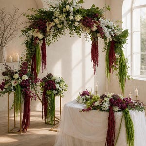 Può includere: Un grande arco floreale con fiori bianchi, bordeaux scuro e verdi con amaranto a cascata, posizionato sopra un tavolo drappeggiato con tessuto bianco. Due composizioni floreali abbinate su supporti dorati fiancheggiano il tavolo con candele.