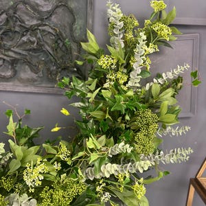 Pode incluir: Uma guirlanda verde exuberante com folhas de eucalipto e hera, acentuada com pequenas flores brancas. A guirlanda &eacute; perfeita para adicionar um toque de natureza a qualquer espa&ccedil;o.