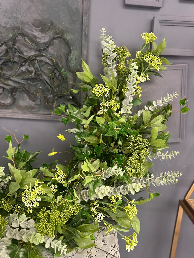 Pode incluir: Uma guirlanda verde exuberante com folhas de eucalipto e hera, acentuada com pequenas flores brancas. A guirlanda &eacute; perfeita para adicionar um toque de natureza a qualquer espa&ccedil;o.