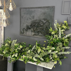 Pode incluir: Uma mesa de m&aacute;rmore cinza com base cinza e uma guirlanda decorativa de folhas verdes e flores brancas. A mesa &eacute; decorada com velas brancas e cinzas.