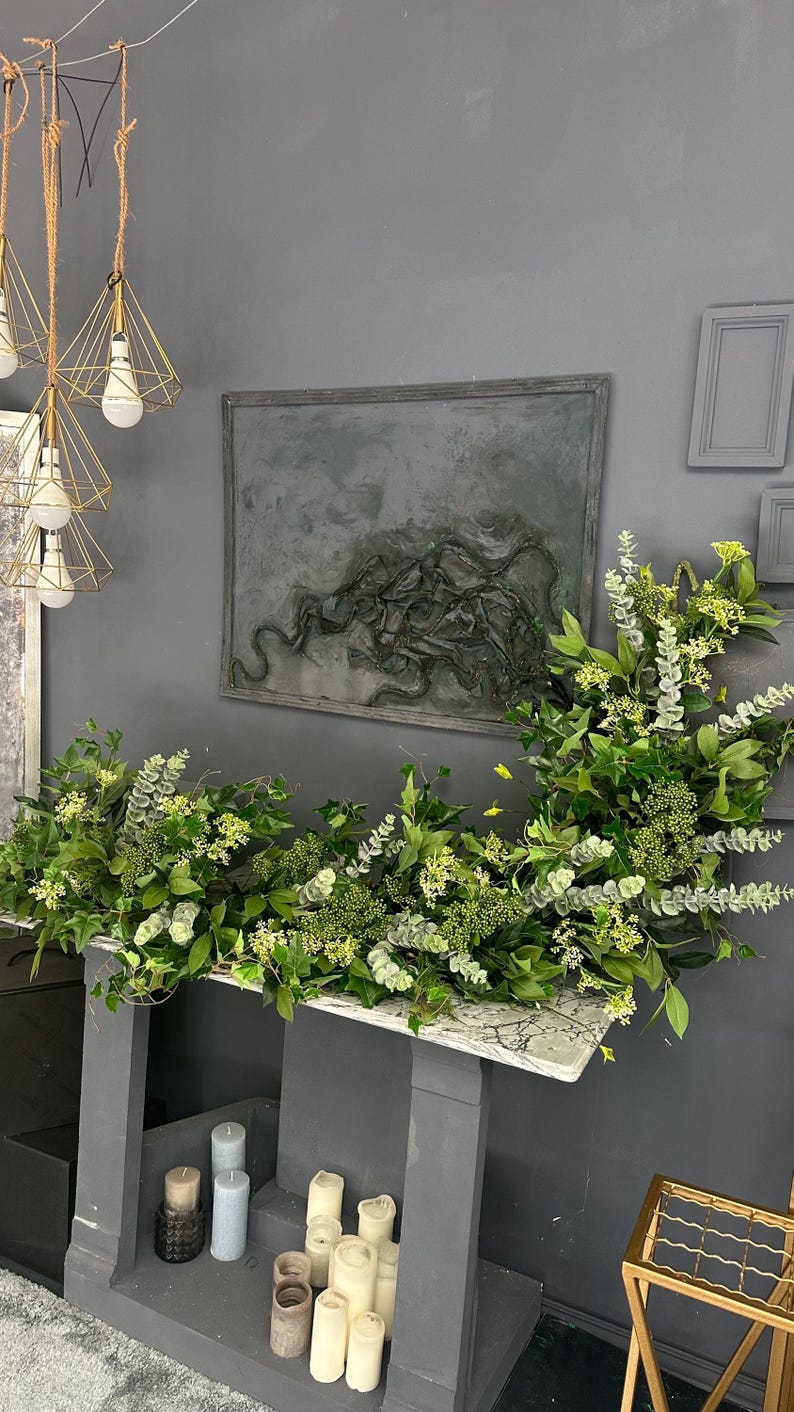 Pode incluir: Uma mesa de m&aacute;rmore cinza com base cinza e uma guirlanda decorativa de folhas verdes e flores brancas. A mesa &eacute; decorada com velas brancas e cinzas.