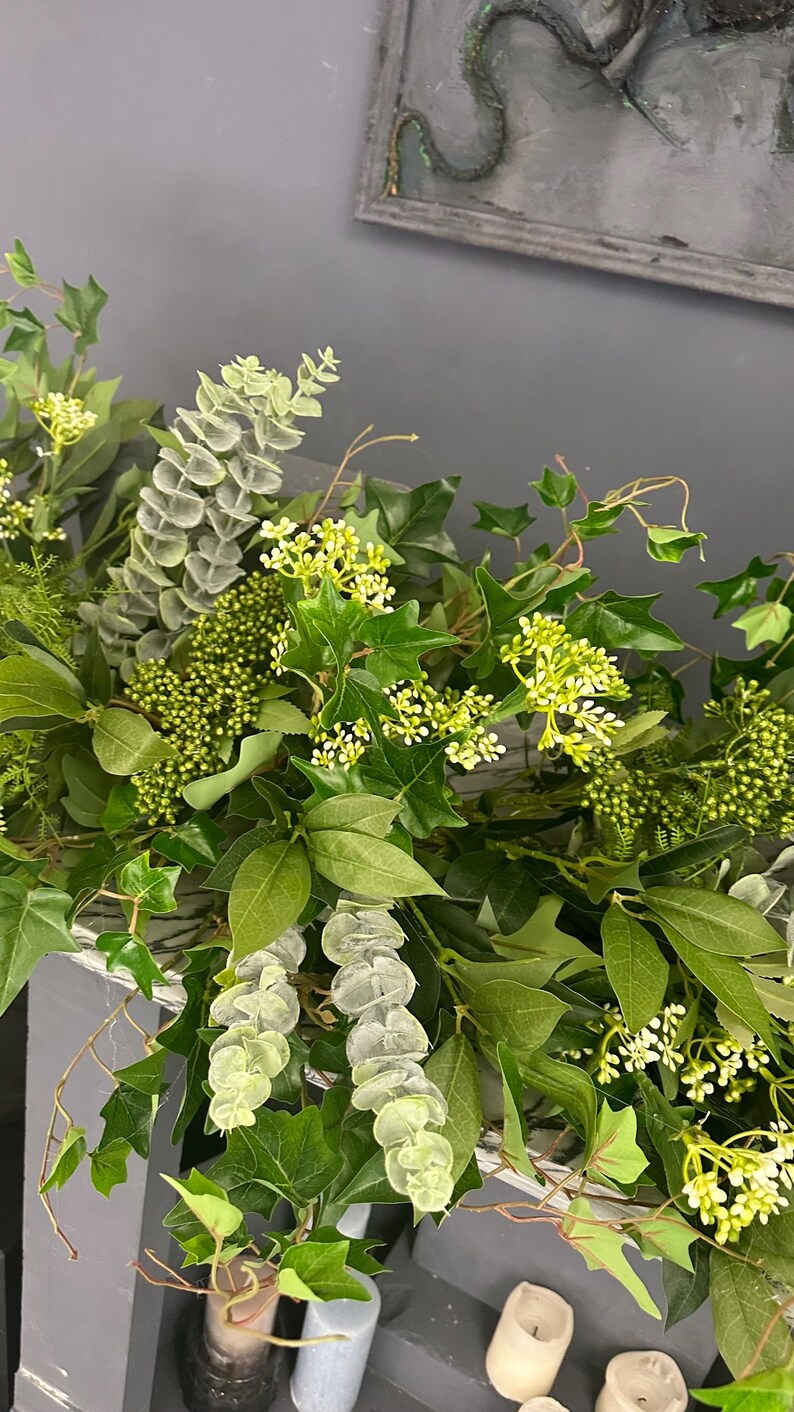 Pode incluir: Uma guirlanda verde com eucalipto, hera e pequenas flores brancas. A guirlanda est&aacute; drapeada sobre uma prateleira com velas.
