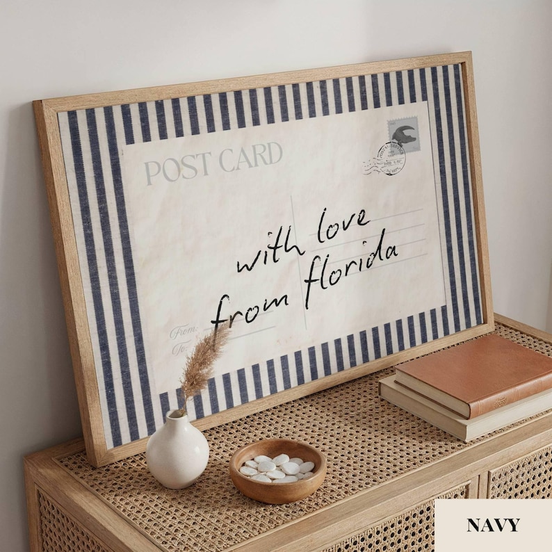 May include: A framed print featuring a vintage postcard design with navy blue and white vertical stripes. The postcard text reads POST CARD and with love from florida in black script. A small white vase and a wooden bowl of white stones sit nearby.
