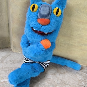 Puede incluir: Un peluche azul con forma de gato con grandes ojos amarillos, una nariz naranja y una boca cosida. Tiene un parche gris a rayas en la cabeza y lleva una banda a rayas blancas y negras. El juguete es suave y esponjoso.