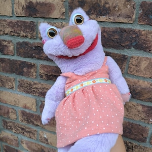 Peut inclure: Marionnette violette avec de grands yeux et une bouche rouge. Elle porte une robe rose à pois blancs et une ceinture à motifs floraux. Le nez de la marionnette est multicolore. L'expression de la marionnette est amicale.