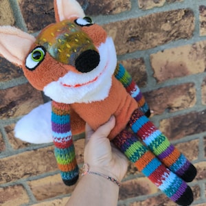 May include: A plush fox toy with orange fur, white accents, and colorful striped legs and arms. The fox has a black nose, green eyes, and a smiling expression. The toy is held against a brick wall.