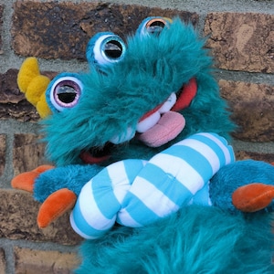 May include: A teal plush monster puppet with shaggy fur, three large glittery eyes, and a yellow horn. The creature has a wide mouth with white teeth, a pink tongue, and orange claws. It is wearing a blue and white striped fabric scarf around its neck.