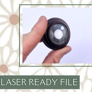 Puede incluir: Primer plano de un pequeño objeto redondo de madera con una lente de vidrio transparente. El objeto tiene un diseño en capas y un círculo blanco en el centro de la lente.