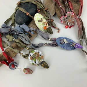 May include: A collection of handmade jewelry featuring colorful ceramic pendants, beads, and charms. The pendants are shaped like hearts, ovals, and circles, and are decorated with floral patterns, geometric designs, and birds. The jewelry is strung on a variety of ribbons and cords, including green, blue, and red.