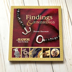 May include: A book titled "Findings & Finishings" by Sharon Bateman, a beadwork how-to book. The cover features a maroon background with jewelry findings and a beaded necklace. The book has a gold border and is on a light wood surface.