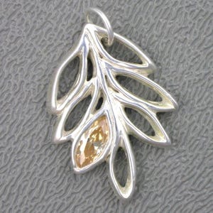 May include: A silver pendant in the shape of a stylized leaf. The pendant features openwork detailing and a central, marquise-cut, amber-colored gemstone. The leaf design has multiple rounded lobes and a loop for a chain.