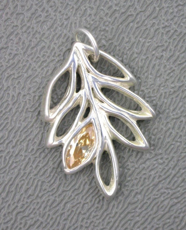 May include: A silver pendant in the shape of a stylized leaf. The pendant features openwork detailing and a central, marquise-cut, amber-colored gemstone. The leaf design has multiple rounded lobes and a loop for a chain.