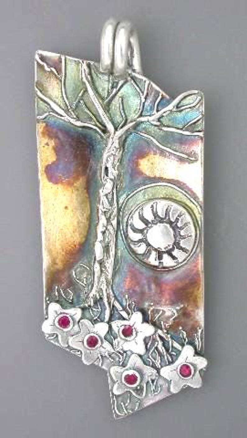 May include: A rectangular silver pendant with a tree design, a sunburst, and flowers with red centers. The pendant has a loop at the top for a chain. The metal has a colorful, iridescent finish.
