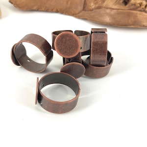 May include: A collection of antique copper-colored adjustable ring blanks. The ring blanks have a rounded band and a flat, circular base for attaching embellishments. The rings are scattered on a white surface, with a piece of wood in the background.