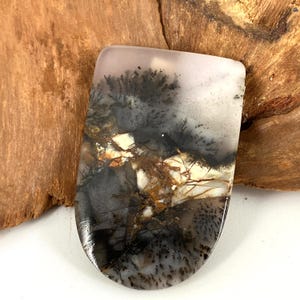 May include: A polished, rectangular gemstone with rounded edges. The stone features a dendritic pattern in shades of black, gray, brown, and white, resembling a landscape or abstract art. The stone is set against a wooden background.