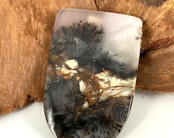 May include: A polished, rectangular gemstone with rounded edges. The stone features a dendritic pattern in shades of black, gray, brown, and white, resembling a landscape or abstract art. The stone is set against a wooden background.