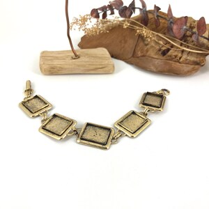 May include: Gold-toned bracelet featuring a series of connected square links. The bracelet has a textured surface and a clasp closure. The background includes decorative wooden elements and dried foliage, creating a rustic aesthetic.