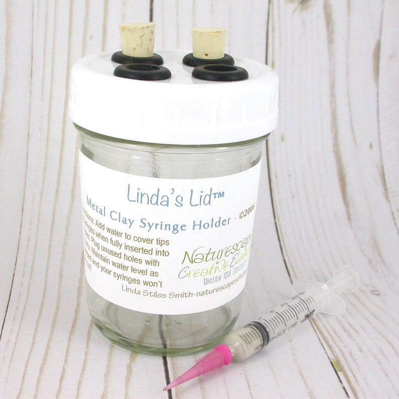 May include: A clear glass jar with a white lid, labeled "Linda's Lid&trade; Metal Clay Syringe Holder." The lid has four holes with cork stoppers and black rubber rings. A syringe with a pink tip rests beside the jar.