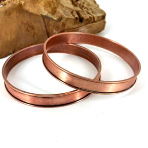 May include: Two copper-colored metal rings with a flat, wide band. The rings are circular and have a slightly textured surface. The background includes a piece of wood. The rings are likely components for jewelry or other crafts.