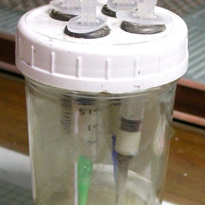 May include: A clear glass jar with a white lid, containing liquid and sediment. Several clear plastic tubes protrude from the lid. Syringes are visible inside the jar. The jar is likely used for scientific or laboratory purposes.