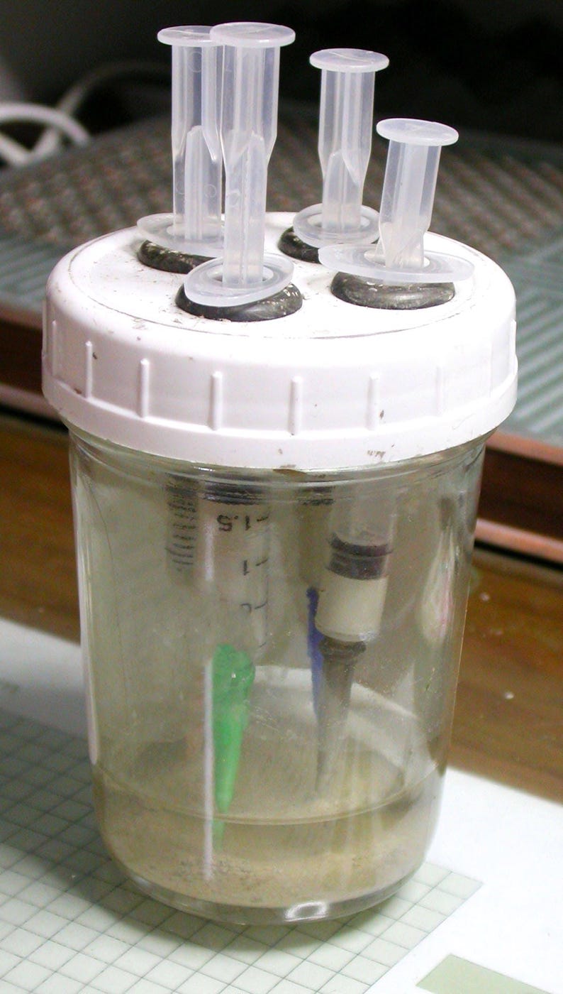 May include: A clear glass jar with a white lid, containing liquid and sediment. Several clear plastic tubes protrude from the lid. Syringes are visible inside the jar. The jar is likely used for scientific or laboratory purposes.