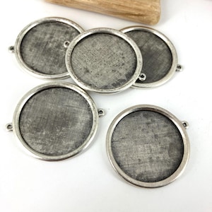 May include: A collection of five silver-toned, round metal bezels with a textured mesh insert. Each bezel has small loops on either side. The bezels are arranged on a white surface, with a piece of wood in the background.