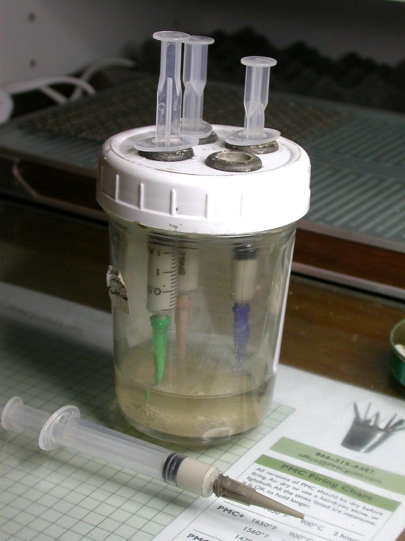 May include: A clear glass jar with a white lid, containing liquid and syringes with colored tips. A syringe lies in front of the jar. A printed chart with text is visible. The jar has four clear plastic tubes on top.