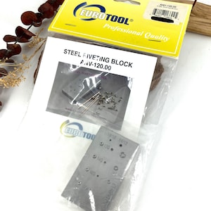 May include: A package containing a steel riveting block with the text "STEEL RIVETING BLOCK ANV-120.00". The block is silver and has various sized holes. The package also includes a smaller block and rivets. The packaging has the "EUROTOOL" logo.