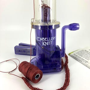 May include: A purple plastic Embellish Knit! machine with a clear top cylinder and a hand crank. A spool of dark red yarn is positioned in front, with a length of knitted cord extending from the machine and wrapping around the spool base.