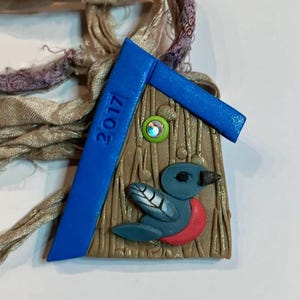 May include: A blue and brown clay ornament shaped like a birdhouse with a bird inside. The ornament has the year 2017 written on it.