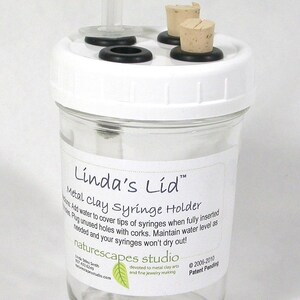 May include: A clear glass jar with a white lid, designed as a metal clay syringe holder. The lid has a clear plastic tube and several corks. The jar has a label with instructions and the text "Linda's Lid".
