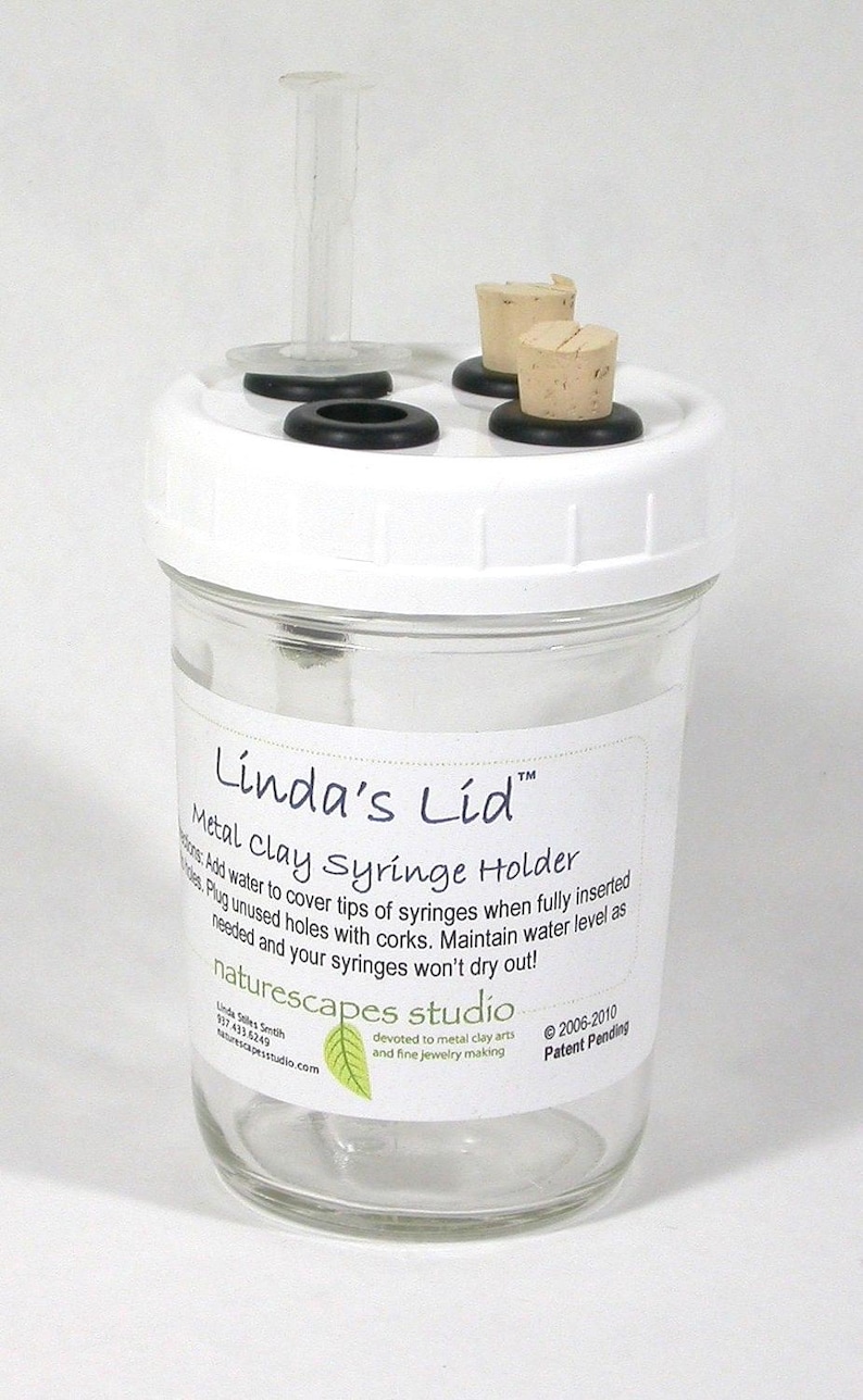 May include: A clear glass jar with a white lid, designed as a metal clay syringe holder. The lid has a clear plastic tube and several corks. The jar has a label with instructions and the text "Linda's Lid".